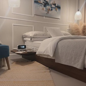 May include: A bedroom scene with a bed dressed in white bedding and a grey quilted throw. A small bedside table holds a digital clock showing 7:00, a cup, and glasses. A blue chair and a rug are also visible.