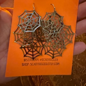 May include: A pair of metallic, silver-toned spiderweb-shaped dangle earrings attached to an orange display card. The card features the text INSTAGRAM: @SCARYRIGGED and SHOP: SCARYRIGGED.ETSY.COM printed in black ink at the bottom.