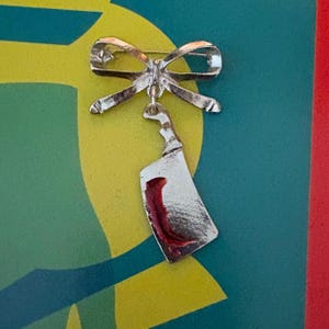 May include: A silver brooch in the shape of a bow with a silver knife hanging from it. The knife has a red stain on the blade.