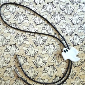 May include: A black bolo tie with a white ghost-shaped slide. The ghost has two black eyes. The background is a repeating pattern of white ghost illustrations on a tan background.