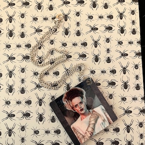 May include: A silver chain necklace with a square pendant featuring a black and white illustration of a woman with bandages on her face and arms. The background of the pendant is a dark, blurry image. The necklace is on a white background with a repeating pattern of black spiders.