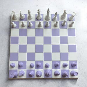 May include: A square chess board featuring a grid of alternating white and light purple squares. The board is set up with a full set of white chess pieces on one side and a full set of matching light purple chess pieces on the opposite side.