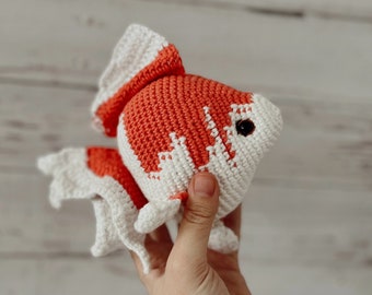 May include: A handmade crochet goldfish in orange and white. The fish has a rounded body, detailed fins, and a small black eye. The fish is held in a hand against a light wood background.