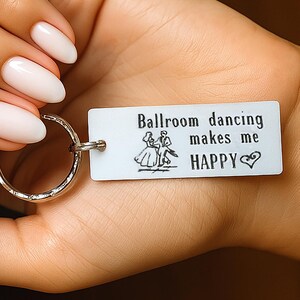 May include: A silver keyring with a rectangular tag that reads "Ballroom dancing makes me HAPPY" with a dancing couple illustration. The keyring is held in a hand with polished nails.