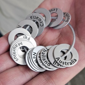 Puede incluir: Una pulsera plateada con colgantes circulares grabados con nombres de países como "Australia", "Tailandia" y "Mauricio". Los colgantes están unidos por un fino alambre, formando un accesorio con temática de viajes.