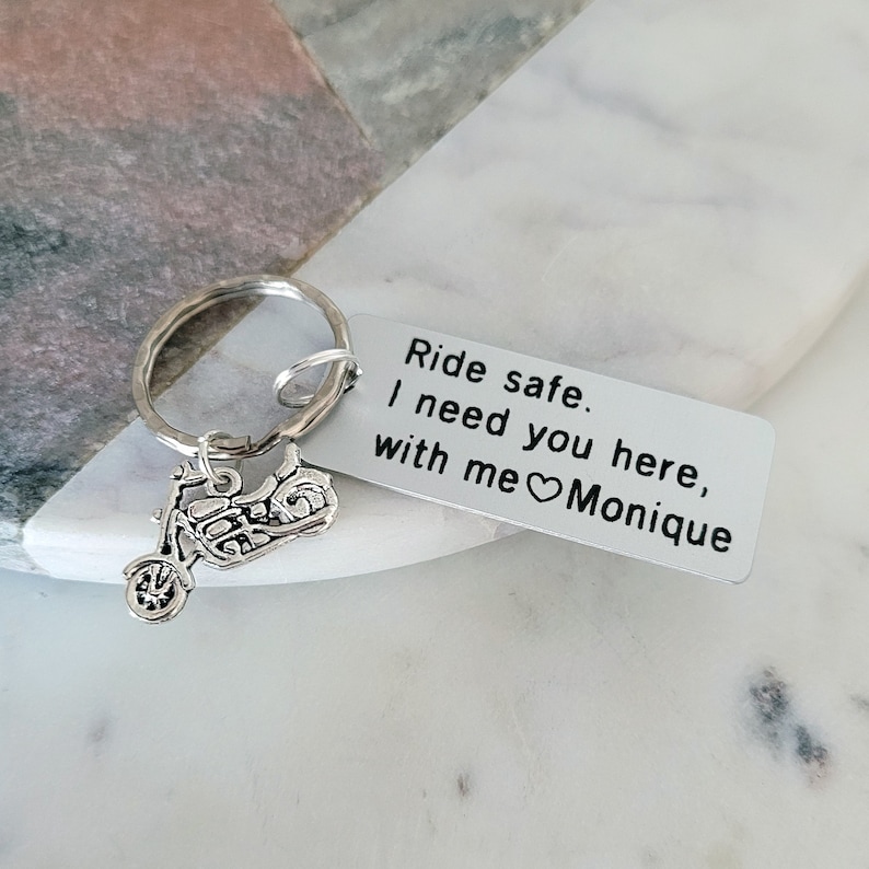 May include: A silver motorcycle keychain with a rectangular tag. The tag reads "Ride safe. I need you here, with me Monique." The keychain is on a marble surface.