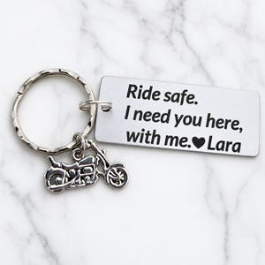 May include: A silver-colored keychain with a motorcycle charm and a rectangular tag. The tag has the text "Ride safe. I need you here, with me. ❤️ Lara". The keychain is on a white marble surface.