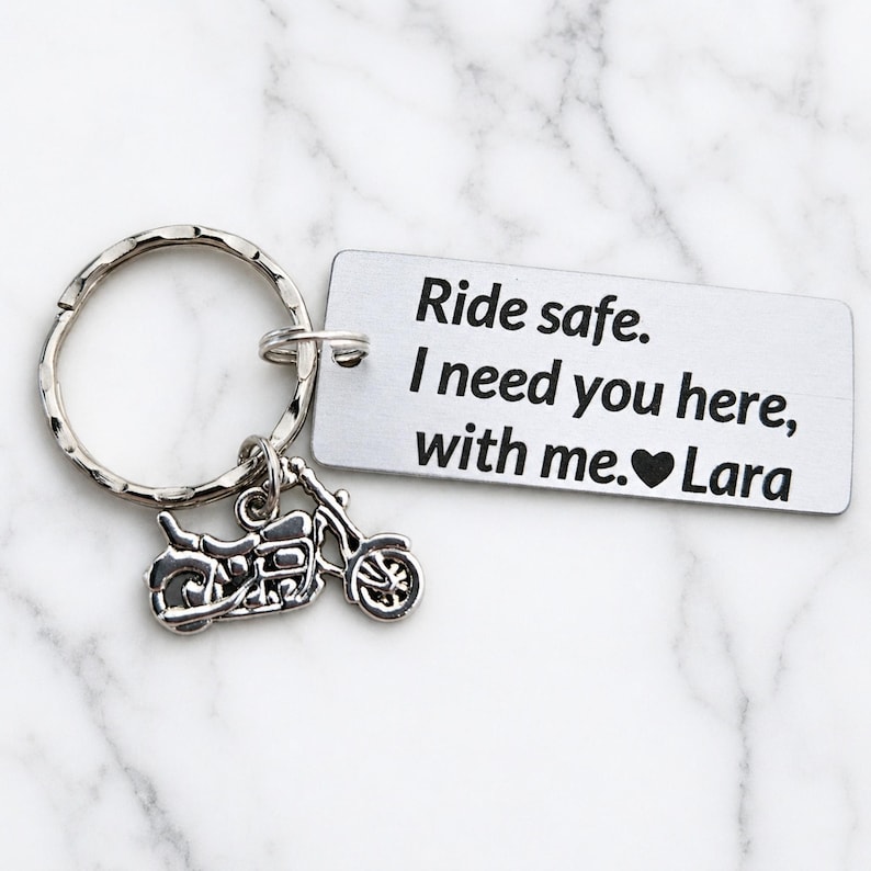 May include: A silver-colored keychain with a motorcycle charm and a rectangular tag. The tag has the text "Ride safe. I need you here, with me. ❤️ Lara". The keychain is on a white marble surface.