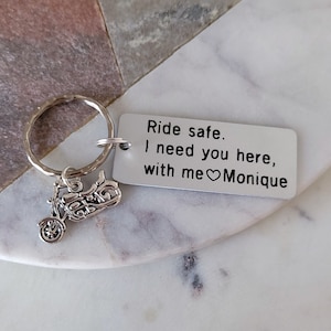 May include: A silver motorcycle keychain with a rectangular tag. The tag reads "Ride safe. I need you here, with me ❤️ Monique." The keychain is on a white and gray marble surface.