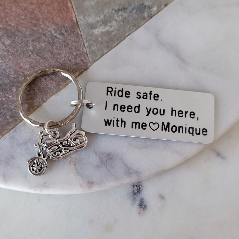 May include: A silver motorcycle keychain with a rectangular tag. The tag reads "Ride safe. I need you here, with me ❤️ Monique." The keychain is on a white and gray marble surface.