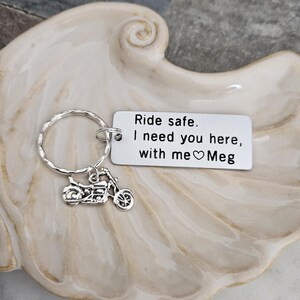 May include: Silver motorcycle keychain with a circular key ring and a rectangular tag. The tag reads "Ride safe. I need you here, with me ♡ Meg". The keychain is displayed on a decorative, off-white, seashell-shaped dish.