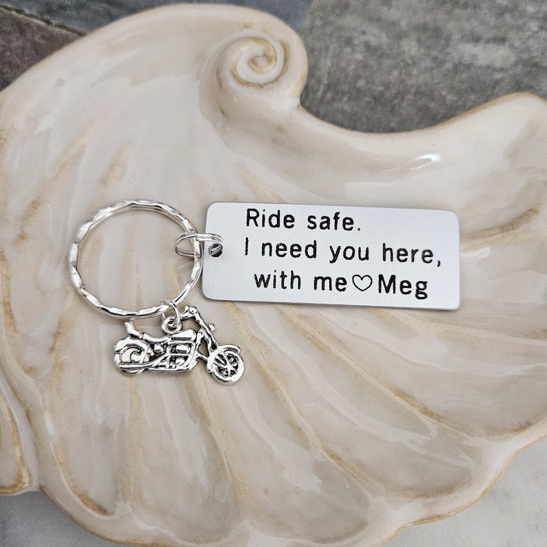 May include: Silver motorcycle keychain with a circular key ring and a rectangular tag. The tag reads "Ride safe. I need you here, with me ♡ Meg". The keychain is displayed on a decorative, off-white, seashell-shaped dish.