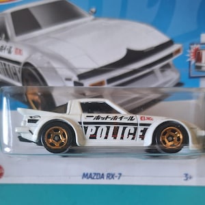 May include: A white Hot Wheels Mazda RX-7 die-cast toy car in its original packaging. The car features gold wheels, a rear spoiler, and the word POLICE printed in black on the side doors, along with Japanese text and the JNC logo on the front fender.