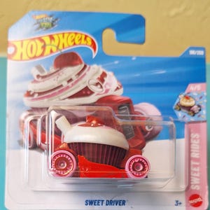 May include: A Hot Wheels Sweet Driver toy car in its original packaging. The vehicle is shaped like a cupcake with a red base, white frosting top, and pink wheels. The blue and white card features the Hot Wheels logo and the text Sweet Rides.