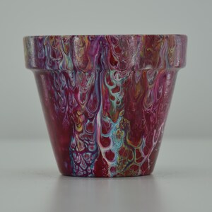 Könnte beinhalten: Ein kleiner Terrakotta-Topf mit einem lebendigen abstrakten Design mit Wirbeln in Pink, Rot, Weiß, Blau und Gold.