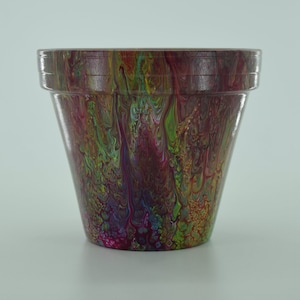 Könnte beinhalten: Ein Terrakotta-Blumentopf mit einem farbenfrohen abstrakten Design in Grün-, Lila-, Rosa- und Goldtönen. Der Topf ist glasiert und hat eine glatte Oberfläche.