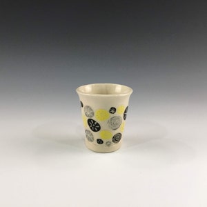 May include: A white ceramic cup with a geometric pattern of yellow and black circles. The cup is a simple, modern design.
