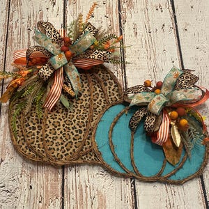 May include: Two decorative pumpkin wreaths, one with a leopard print and the other with a turquoise blue fabric. Both pumpkins are adorned with fall-themed ribbons and floral accents.