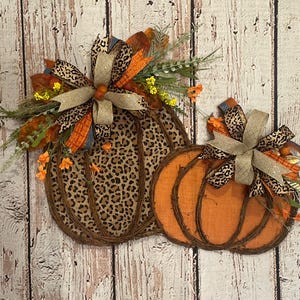 May include: A fall-themed door wreath featuring two pumpkins. The larger pumpkin is covered in a brown and black leopard print fabric. The smaller pumpkin is a solid orange color. Both pumpkins have a brown twig-like texture and are decorated with a brown and black leopard print bow.