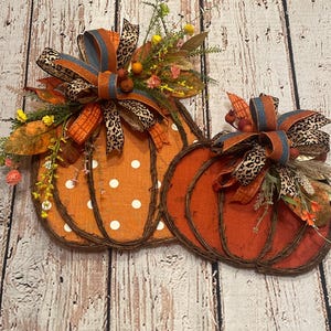 May include: Two orange and brown pumpkins with white polka dots and a leopard print bow. The pumpkins are made of woven twigs and have a rustic look.