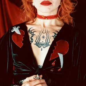 May include: A black velvet top with a plunging neckline, adorned with two red broken heart appliqués and silver accents. The person in the image has red hair, red lipstick, and a red choker. The person has tattoos on their chest and arms.