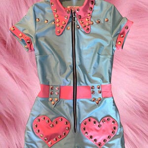 Puede incluir: Un mono corto de manga corta de color azul claro con cuello rosa y detalles rosas. El mono tiene una cremallera y está decorado con tachuelas plateadas. El mono tiene dos parches en forma de corazón rosa en la parte delantera.