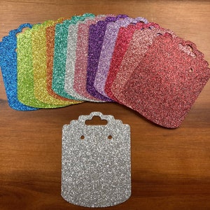 May include: Assortment of colorful glitter earring display cards. The cards are square shaped with a hole at the top for hanging. The cards are made of thick cardboard and have a glitter finish. The cards are available in a variety of colors, including blue, green, yellow, orange, red, pink, purple, and silver.