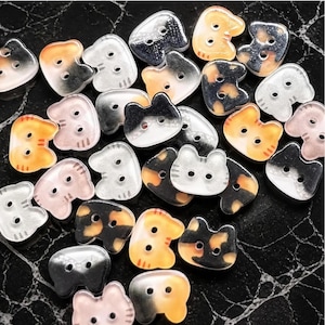 May include: A collection of small, two-hole buttons shaped like cat heads. The buttons feature various color patterns including orange, black, white, and grey, with some showing tabby stripes or calico-style patches on a smooth, semi-translucent surface.