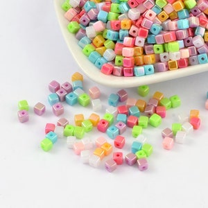 May include: A bowl filled with iridescent pastel colored square beads. The beads are scattered on a white surface.