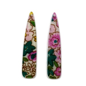 Puede incluir: Dos pinzas para el pelo largas y estrechas con un patrón floral. Las pinzas son blancas con flores rosas, verdes y marrones.