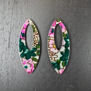 Puede incluir: Dos pendientes ovalados con un estampado floral. Los pendientes presentan un diseño floral rosa y blanco con hojas verdes. Los bordes están delineados en rosa. Los pendientes son de material brillante.