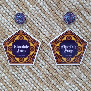 May include: A pair of purple glitter earrings with a silver charm featuring the Chocolate Frogs logo from the Harry Potter series. The logo is a pentagon with a gold border and the words "Chocolate Frogs" in white text.