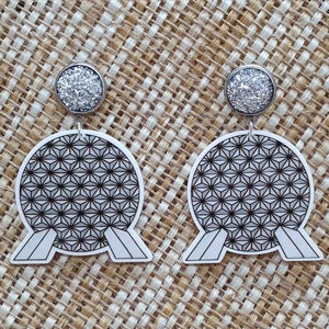 May include: A pair of white dangle earrings with a black and white geometric design of the Spaceship Earth attraction at Epcot. The earrings have silver glitter tops.