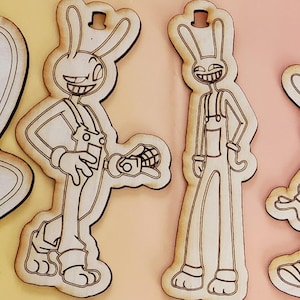 May include: Wooden cutouts of a cartoon rabbit character in overalls. The rabbit has a wide grin and is holding a grenade. The cutouts are light brown with dark brown outlines and a hole at the top for hanging.