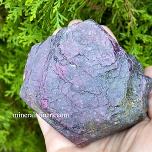 May include: A large, rough, purple ruby crystal with a dark purple hue and a slightly green tint. The crystal has a natural, unpolished surface.