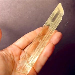 May include: A pale yellow, translucent, elongated crystal with visible internal striations.  The crystal is roughly rectangular with slightly irregular edges and a pointed top.  The color is consistent throughout, showing a slight variation in tone due to light refraction. The crystal displays a natural, unpolished appearance.  The website mineralminers.com is visible in the image.