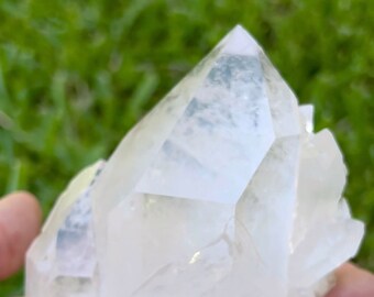 May include: A large, clear quartz crystal cluster with multiple points. The crystal is translucent and has a slight milky hue. The crystal is being held in a hand.