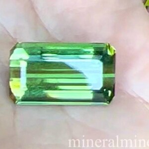 May include: A green gemstone with a rectangular shape and a faceted cut. The gemstone has a light green color and a slightly yellow hue. The gemstone is cut in a step cut style, with a series of parallel lines running across the surface. The gemstone is reflecting light and has a shiny appearance. The text 'mineralminers.com' is visible in the lower right corner of the image.