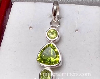 May include: A silver pendant with three green peridot gemstones. The pendant is in a white box with a red border. The text "mineralminers.com fluorescent light" is visible at the bottom of the image.