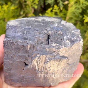 May include: A large, rough-hewn, metallic gray mineral specimen. The surface has a textured, layered appearance with visible crystalline structures. The mineral is held in a hand, with a blurred green background. The website address "mineralminers.com" is visible.