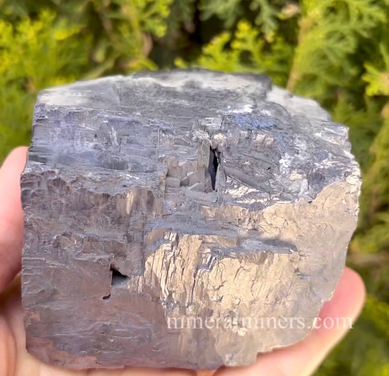 May include: A large, rough-hewn, metallic gray mineral specimen. The surface has a textured, layered appearance with visible crystalline structures. The mineral is held in a hand, with a blurred green background. The website address "mineralminers.com" is visible.