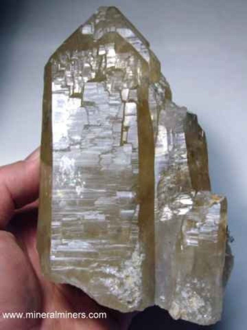 May include: A large, translucent, light-brown crystal with a faceted, geometric structure. The crystal has a pointed top and a rough, textured surface. The image is of a mineral specimen, likely for display or collection.