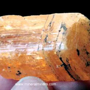 May include: A large, orange-brown, translucent crystal with striations and black inclusions. The crystal is a natural mineral specimen.