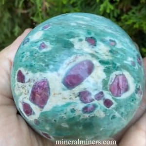 May include: A spherical gemstone with a teal and white base color, speckled with vibrant, deep red inclusions. The polished stone is held in a hand, with the website address mineralminers.com visible. The background is blurred green foliage.
