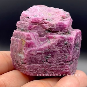 Può includere: Un grande cristallo di rubino rosa con una superficie ruvida e sfaccettata. Il cristallo ha un colore rosa intenso ed è circondato da cristalli neri più piccoli.
