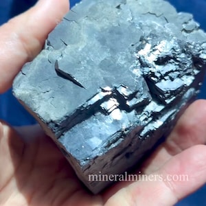 May include: A dark gray mineral specimen with a layered, crystalline structure. The surface has a metallic sheen and visible fractures. The image includes the text "mineralminers.com".