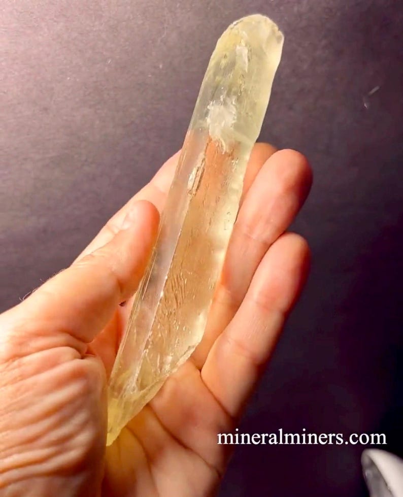 May include: A large, clear, yellow crystal with striations. The crystal is about 10 inches long and is held in a hand.