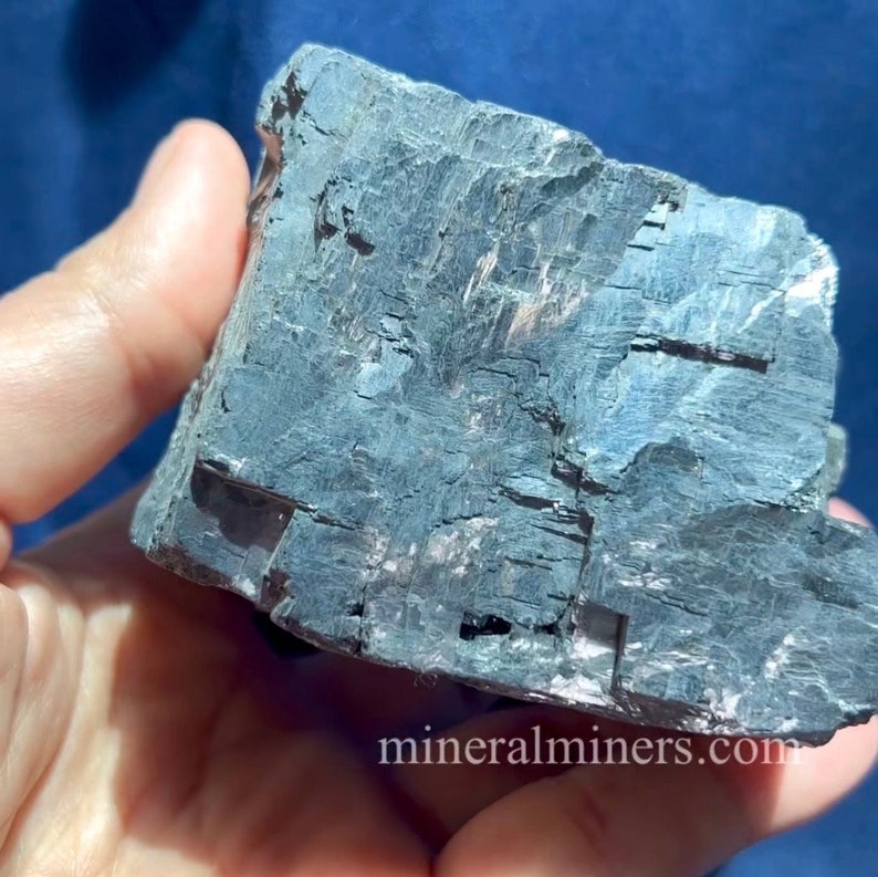 May include: A dark gray mineral specimen with a layered, textured surface. The mineral has a metallic sheen and is held in a hand. The website address mineralminers.com is visible in the image.