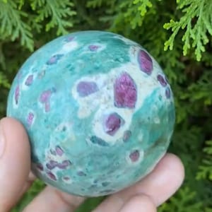 Op de afbeelding: Een ronde, gepolijste edelsteen met een turquoise basiskleur en onregelmatige vlekken van diep roze. De steen heeft een glad oppervlak en wordt tegen een achtergrond van groen gebladerte gehouden. De website "mineralminers.com" is zichtbaar.