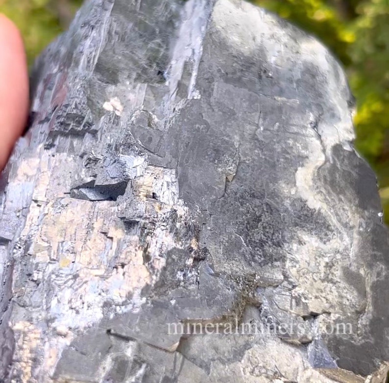 May include: A close-up of a dark gray mineral specimen with a rough, textured surface. The rock has areas of silver and white, with visible crystalline structures. The image is well-lit, highlighting the mineral's natural details.